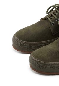 Chaussures en daim vert avec un bout rond, semelle plate en caoutchouc texturé et accents bruns contrastants sur la semelle extérieure et les coutures.