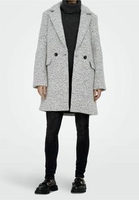 Osoba v lehkém šedém texturovaném kabátu přes tmavý rolák, černých skinny džínách a černých mokasínech se zlatými detaily.