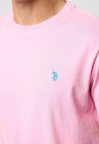 Šviesiai rožinė medvilninė marškinėlių su mažuoju žaliu siuvinėtą logotipu kairėje krūtinės pusėje. Apvalus iškirpimas ir trumpomis rankovėmis.