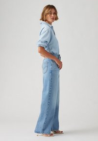 Woman standing sideways wearing rolled-up light blue denim shirt, high-waist wide-leg blue jeans, and beige heeled sandals on plain background.
