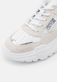 Vita och beige sneaker med perforerad tå, vita snören och "Versace Jeans Couture"-logotyp på sidan.