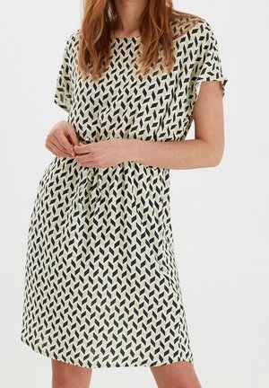 Femme portant une robe à manches courtes avec un motif géométrique chevron noir et beige, ajustant une ceinture nouée à la taille sur un fond uni.