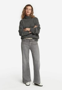 Grauer gestrickter Rollkragenpullover mit gerippter Struktur, kombiniert mit taillierten, ausgestellten grauen Jeans und weißen Schuhen. Einfaches Design, keine Muster.