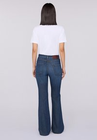 Jeans acampanados azul con un lavado oscuro, que incluyen una cintura alta, bolsillos traseros y un parche de cuero marrón en la cintura, combinados con una camiseta blanca.