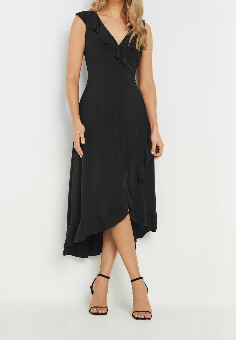 Femme portant une robe portefeuille noire sans manches avec un volant et des escarpins noirs à bride à la cheville, debout sur un fond blanc.