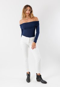 Donna con capelli mossi che indossa un top blu navy a spalle scoperte, pantaloni bianchi, stivaletti neri alla caviglia e una delicata collana d'oro, in piedi di fronte a uno sfondo bianco.