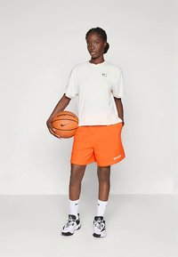 Conjunto de baloncesto que incluye una camiseta blanca de manga corta, pantalones cortos naranjas brillantes con la inscripción "WNBA" y zapatillas negras y blancas, sosteniendo un balón de baloncesto.