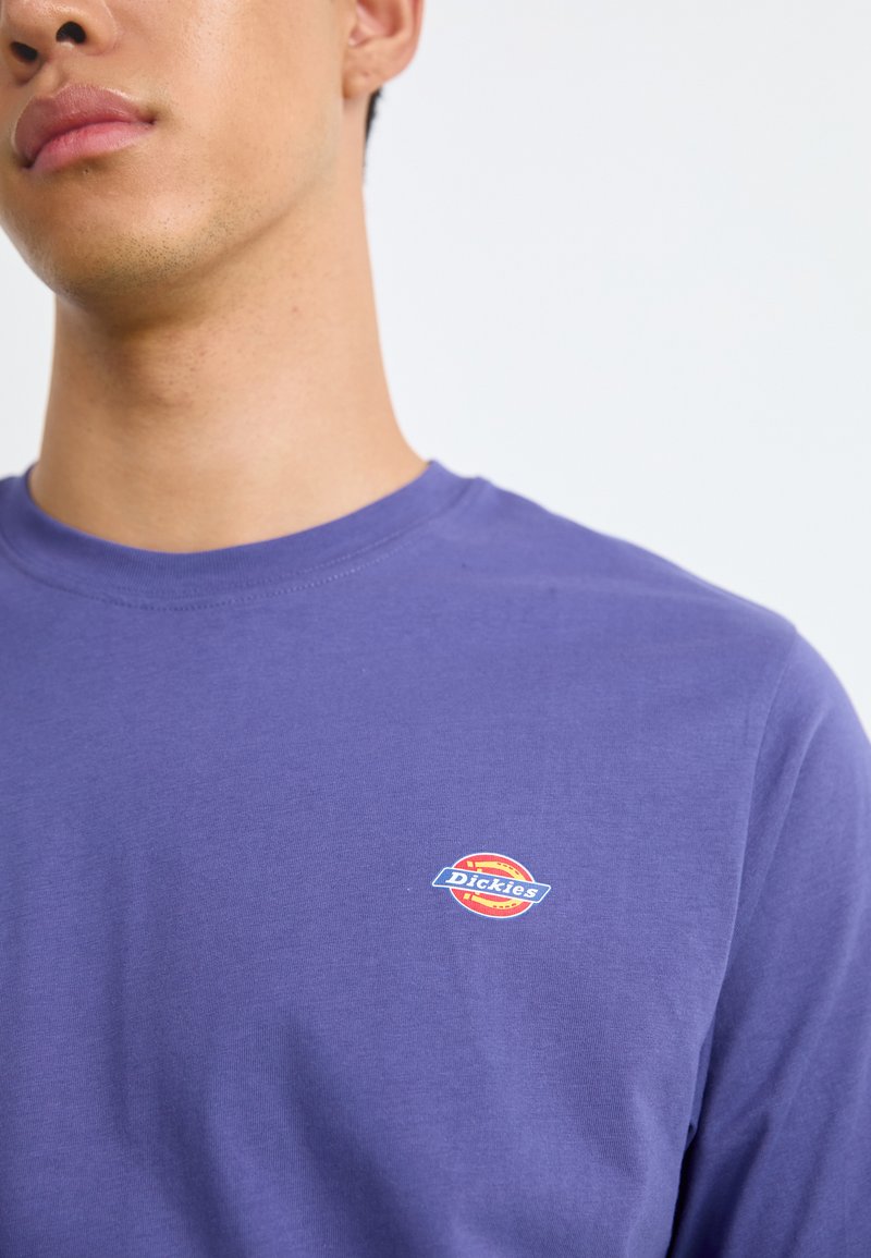 Lila Langarm-T-Shirt aus Baumwolle, mit einem kleinen, mehrfarbigen Dickies-Logo auf der linken Brust. Glatte Textur.