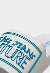 Άσπρο σανδάλι με λευκή σόλα και μπλε περίγραμμα του λογότυπου "Versace Jeans Couture" στο λουράκι, σε ανοιχτό φόντο.