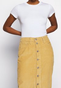 Vit kortärmad åtsittande t-shirt kombinerad med en knälång gul kordkjol som har knapprad fram och sidfickor.