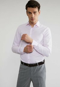 Jasnoróżowa koszula z długim rękawem o teksturowanym wykończeniu, z zapięciem na guziki z przodu i szpiczastym kołnierzykiem, noszona z szarymi spodniami w kratę i czarnym paskiem.