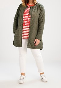 Olive green quilted jacket with a rounded collar, paired with a red striped shirt, white pants, and white slip-on shoes.