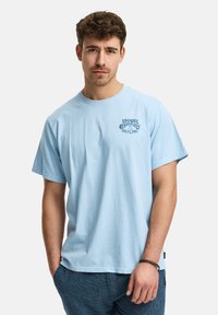 Jeune homme aux cheveux bouclés courts portant un T-shirt bleu clair avec le texte "Votre guérison énergétique" et un pantalon bleu foncé, se tenant avec une main dans sa poche.