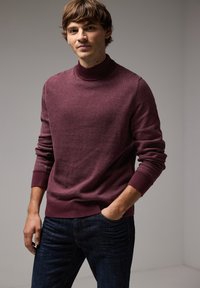 Burgundin värinen korkeakauluksinen neule pehmeästä materiaalista, jossa on ribbireunat hihansuissa ja helmassa, yhdistettynä tummansinisiin farkkuihin rentoa ilmettä varten.