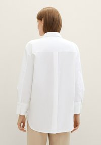 Chemise blanche à boutons avec un empiècement au dos et des manches longues, présentant un ourlet droit et une texture subtile. Portée par-dessus un pantalon crème.