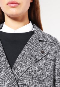 Cappotto grigio in tweed con colletto a punte, caratterizzato da una texture sottile e un accento di bottone nero. Indossato sopra una maglia nera con colletto bianco.