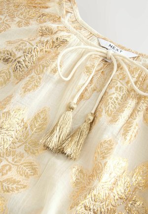 Cream blouse with gold leaf pattern, lightweight fabric, ties at neckline with two gold tassels, and subtle sheen.