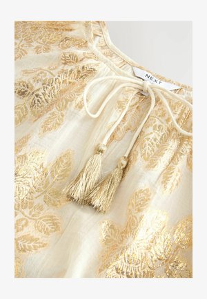 Blusa crema con patrón de hojas doradas, tejido ligero, lazos en el escote con dos borlas doradas y un sutil brillo.