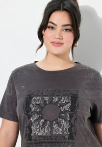 Ubleget sort bomulds t-shirt med et grafisk print, der viser teksten "PARADISE COVE" og et solmotiv, tekstureret med et slidt udseende.
