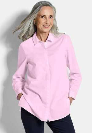 Vrouw met grijs haar draagt een lichtroze blouse met knopen en donkere broek, staat met handen in de zakken en glimlacht tegen een effen achtergrond.