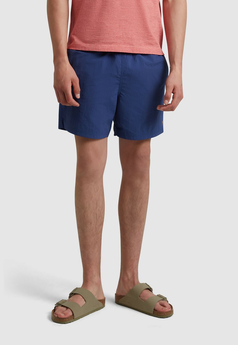 Costume da bagno blu navy in tessuto leggero. Indossato con una maglietta rossa e sandali verdi oliva, con suola piatta e cinturini regolabili.
