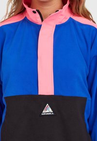 Femme portant une veste polaire bleue et noire avec des accents roses et un logo triangulaire O'Neill sur la poitrine.