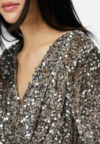 Smėlio spalvos blizgančiais siūtais viršus su giliais V formos iškirpte ir tekstūruota medžiaga, papuošta blizgančiais sidabriniais blizgučiais įvairių dydžių, suteikiančių vizualinį įdomumą.