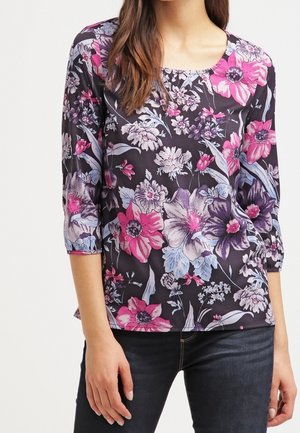 Kvinde iført sort top med pink, lilla og hvidt blomsterprint og trekvartlange ærmer, kombineret med mørke jeans.