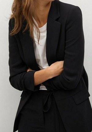 Femme portant un blazer noir et un pantalon noir avec un t-shirt blanc, bras croisés, debout devant un fond clair uni.