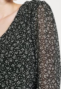 Svart tyg med vita prickar, som har en slät, genomskinlig textur och en V-ringad design. Ärmen har en mjuk fall.