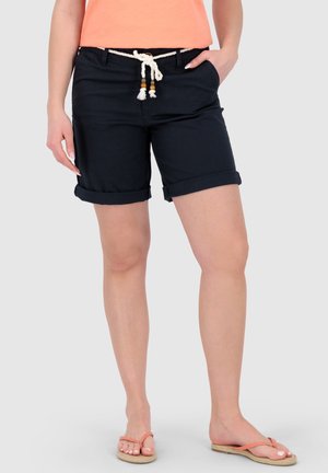 Frau trägt schwarze Shorts mit umgeschlagenem Saum und weißem Kordelgürtel, pfirsichfarbenes Top und korallenfarbene Flip-Flops, steht vor einem hellen Hintergrund.