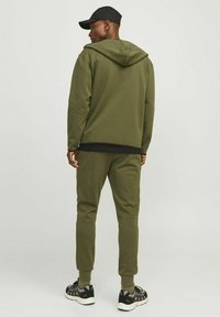 Sweat à capuche vert olive et jogging en tissu doux, avec une coupe décontractée, des poignets côtelés et une capuche. Associé à des chaussures de sport noires.