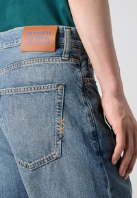 Blue denim jeans with a brown leather patch labeled "SCOTCH & SODA." Notable multicolored stitching on the back pocket.