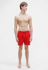 Κόκκινα μαγιό σορτς με ελαστική μέση και κορδόνι, με κομψό σχεδιασμό. Το μοντέλο στέκεται ξυπόλητο μπροστά σε απλό φόντο.