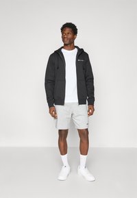 Schwarze Zip-Hoodie, weißes Crewneck-T-Shirt, graue Shorts, weiße Sneaker und weiße Socken. Enthält Logodetails auf den Kleidungsstücken.