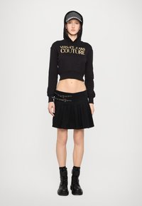 Svart kort hoodie med guldtext, kombinerad med en svart plisserad kjol med spänneaccent och svarta ankelboots.