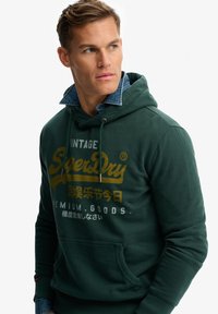 Groene hoodie gemaakt van katoen, met een voorvak in kangoeroestijl, capuchon met trekkoorden en bedrukt merklogo in goud en wit. Denim kraag zichtbaar.