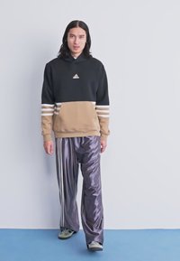 Mlad oseba z dolgimi temnimi lasmi, oblečena v črno-bež Adidas kapuco in sijoče ohlapne sive Adidas trenirke, stoji ob beli steni.
