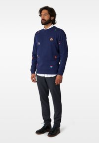 Mornarsko modra trenerka z različnimi barvnimi vezeninami, okrogla ovratnika, ribana manšeta in rob; v kombinaciji s sivimi teksturiranimi hlačami in črnimi čevlji.