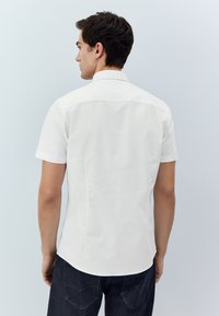 Chemise blanche à manches courtes avec col classique, texture lisse et coupe ajustée, vue de dos avec un jean foncé.