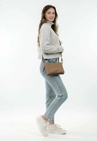Borsa a tracolla in pelle marrone con una texture liscia, dotata di chiusura a zip e tracolla regolabile. Il modello indossa un maglione a coste grigio chiaro e jeans.