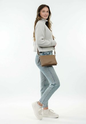 Brown leather crossbody bag with a smooth texture, featuring a zip closure and adjustable strap. Model wears a light gray ribbed sweater and jeans.