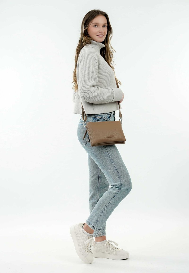 Borsa a tracolla in pelle marrone con una texture liscia, dotata di chiusura a zip e tracolla regolabile. Il modello indossa un maglione a coste grigio chiaro e jeans.