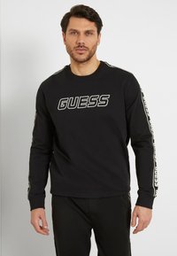 Felpa nera a maniche lunghe con logo "GUESS" stampato sul davanti, caratterizzata da dettagli decorati lungo le maniche e un colletto tondo.