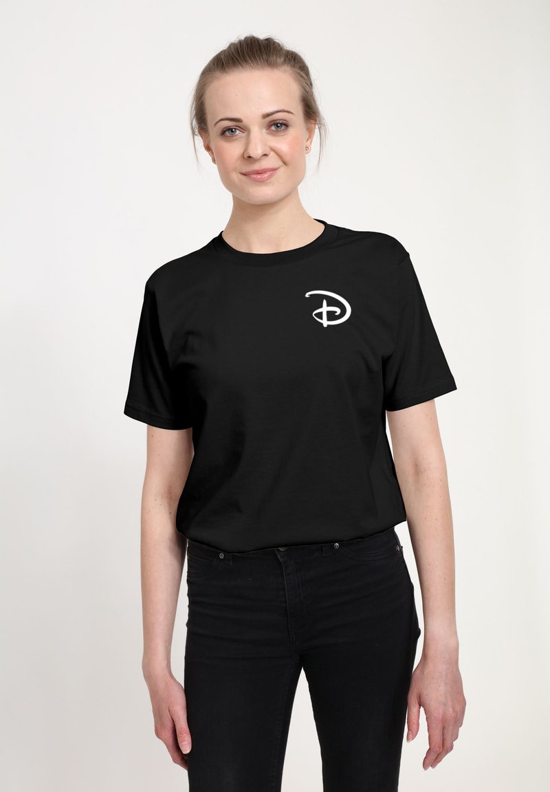 Kvinde med lyst hår iført en sort t-shirt med et hvidt stiliseret "D" logo på brystet, kombineret med sorte bukser, stående mod en hvid baggrund.