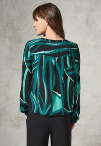 Blusa nera fluente con motivi astratti di foglie verdi e bianche. Presenta un colletto arricciato e maniche lunghe con polsini elasticizzati.