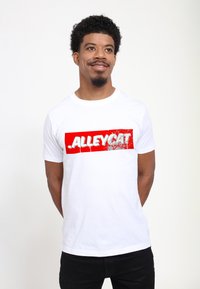 Bílý bavlněný tričko s výraznou červenou grafikou a textem "ALLEYCAT" ve zničeném písmu, nošené s černými kalhotami.