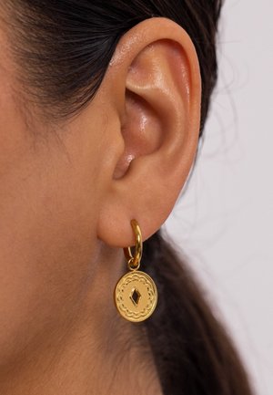 Gros plan sur l'oreille d'une personne portant une boucle d'oreille en or en forme de créole avec un pendentif rond gravé présentant une découpe en forme de diamant.