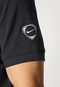 Sort polo-shirt med struktureret stof, korte ærmer, og et sølv- og hvidt Nike-logo broderet på venstre ærme.