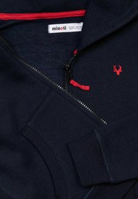 Felpa con zip di colore blu navy, dalla superficie testurizzata, dotata di una linguetta rossa per la zip e un logo di cervo ricamato. Presenta polsini a coste e un rivestimento interno morbido.